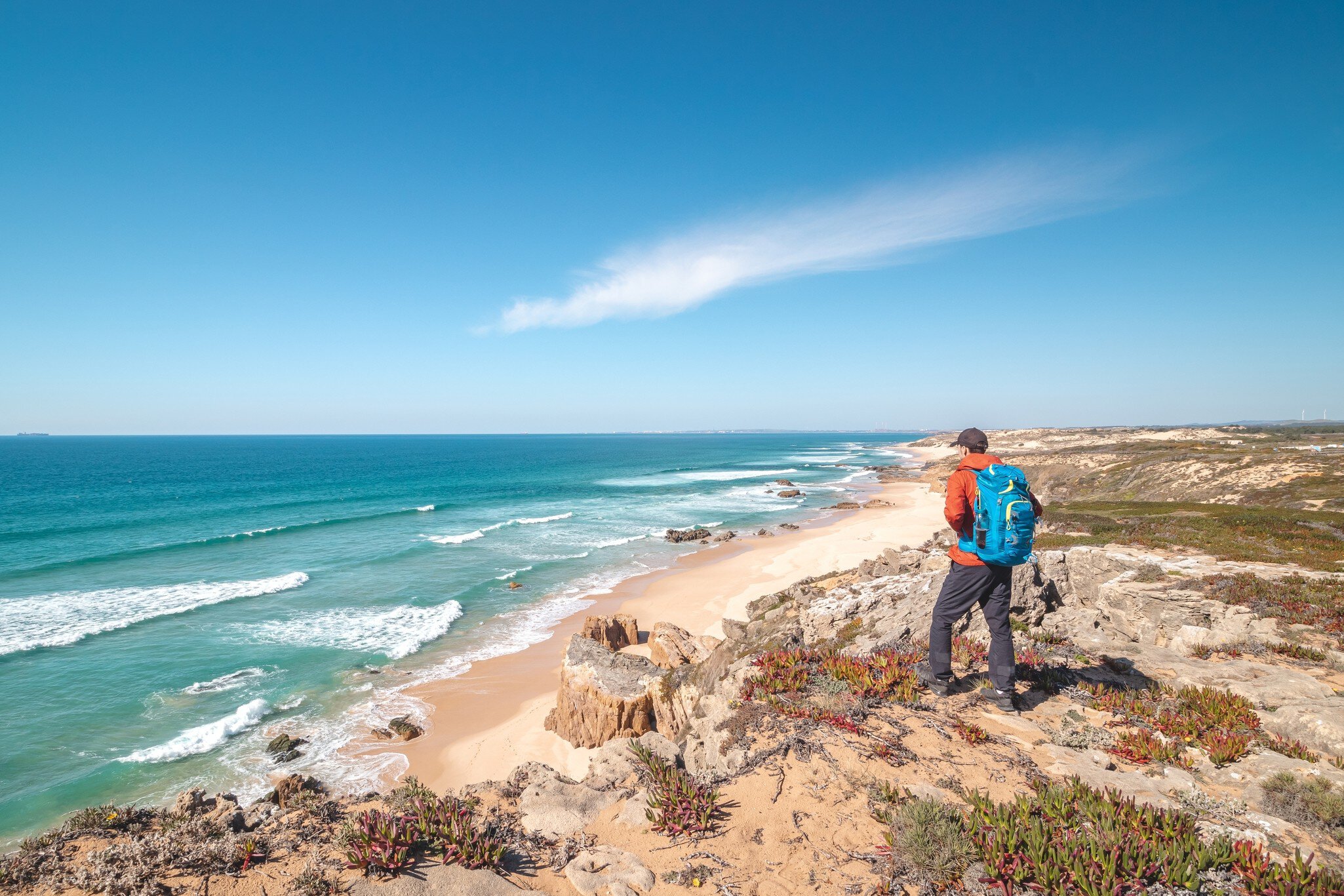 A hiker with a pack is seen from behind standing on dunes and looking out at the waves lapping a beach.