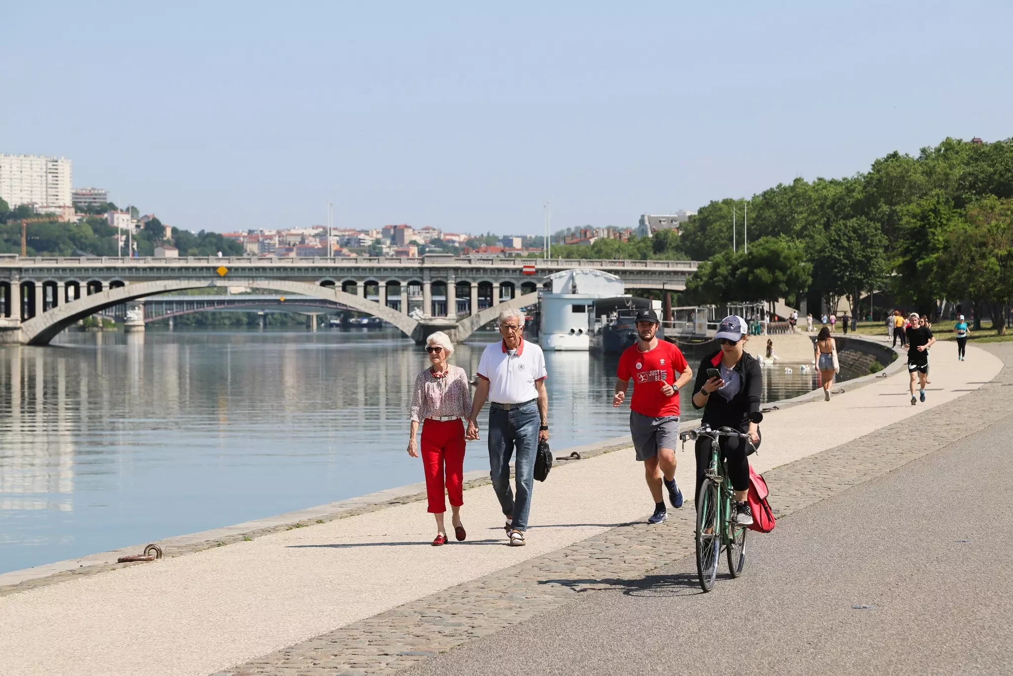 People walk, jog and ride bicycles along the bank of a river in a city.