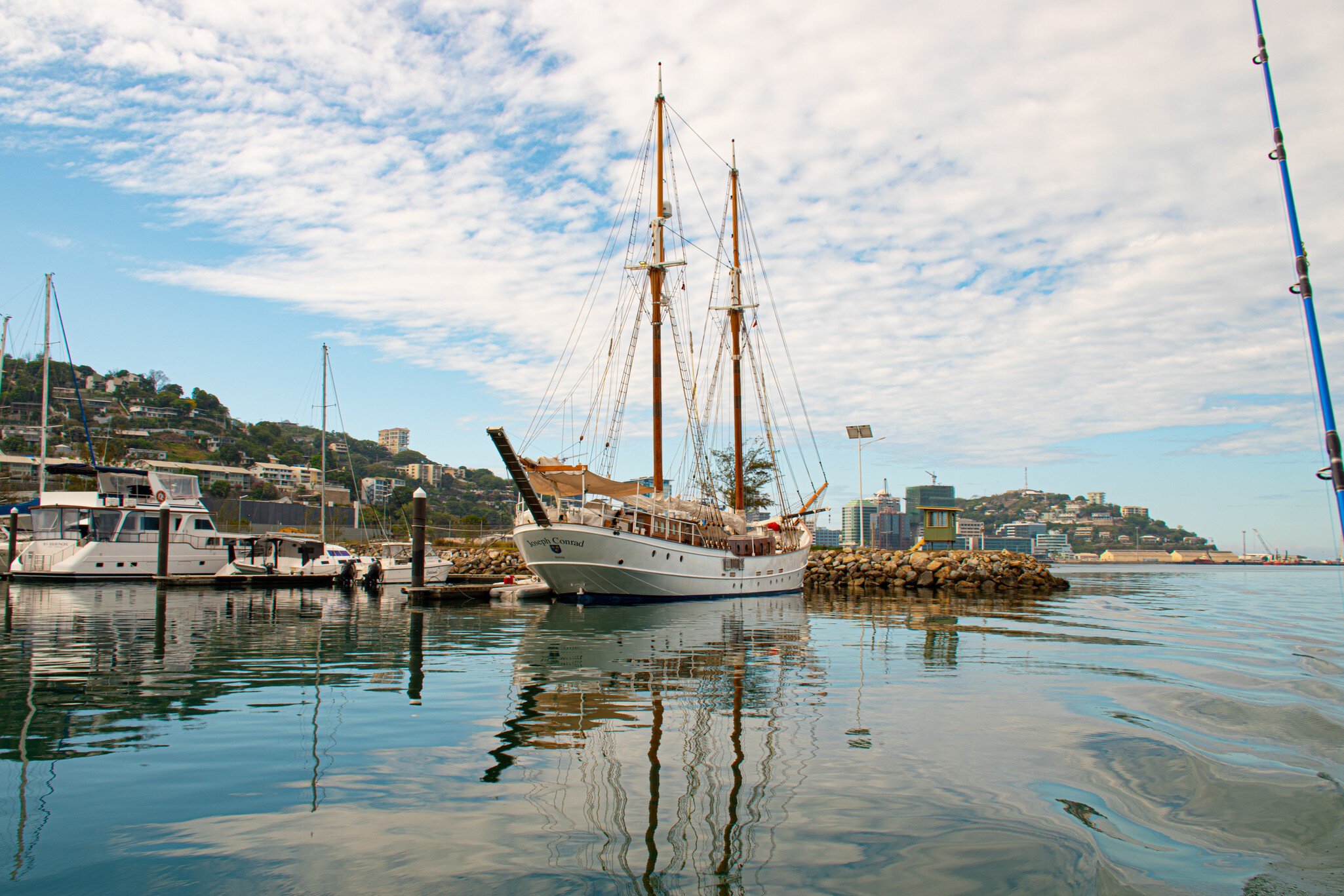 Two-masted schooner Joseph Conrad moored by a breakwater in Port Moresby, Papua New Guinea, with hillside homes behind