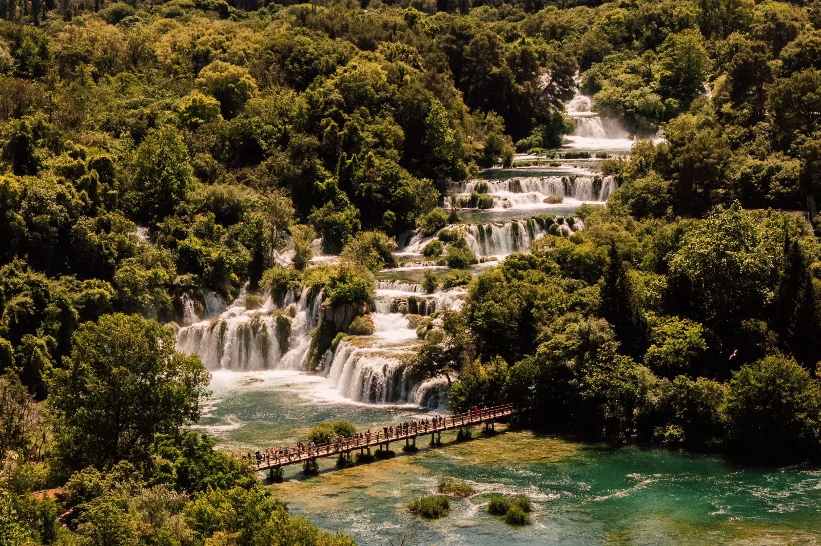 People swimming in a river by a waterfall surrounded by green trees on a sunny day