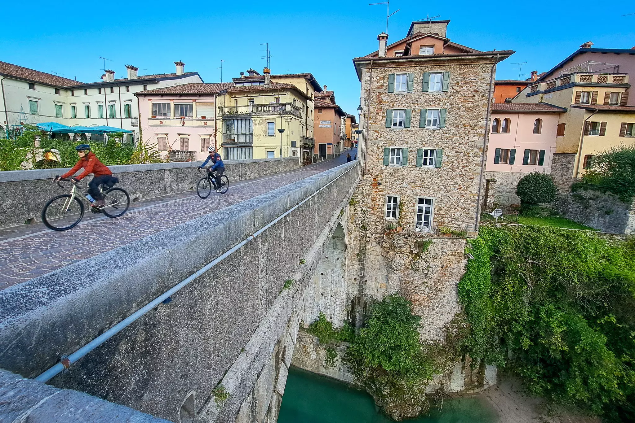 Two bikepacking cyclists ride over cobblestones on a bridge near an old town.