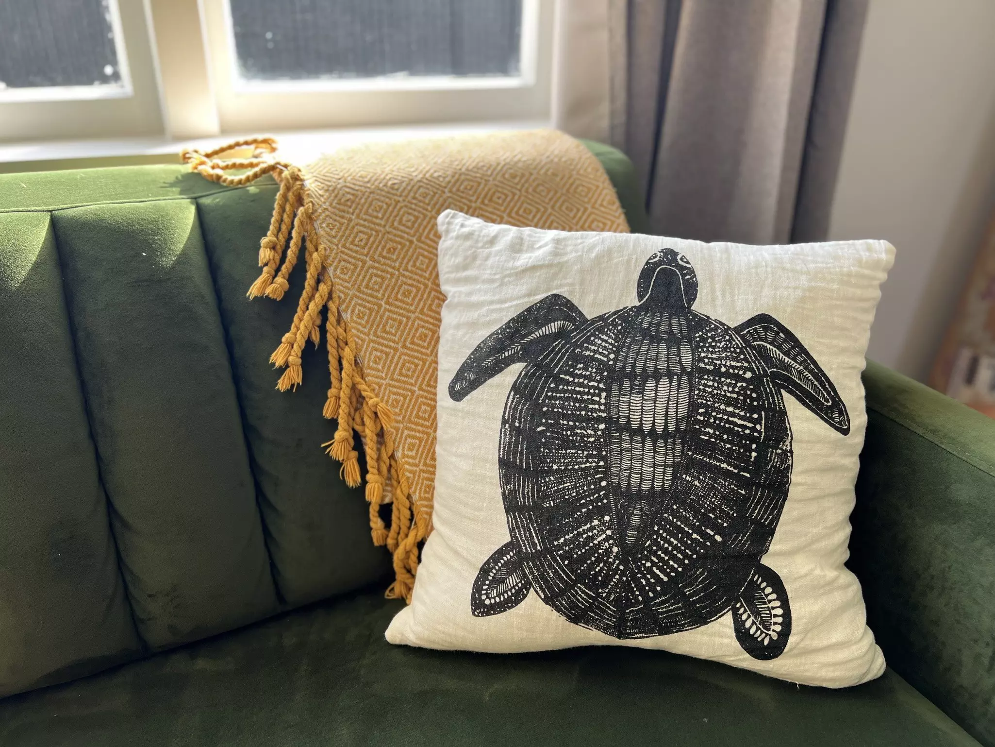 A hand-printed white sofa pillowcase featuring a black ink image of a tortoise on a green sofa with a yellow blanket behind it and sun streaming in from a window beyond.