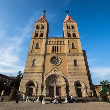 A twin-towered stone church with red spires and a rose window, brides in white gowns gathered on the plaza below.