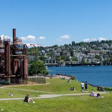 People sitting on the grass on the lakeshore at a park. Buildings and trees rise from the hill across the lake.