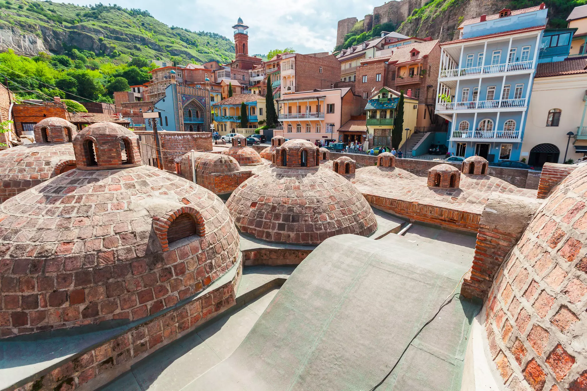 Domed red-brick roofs in the foreground with colorful buildings and hills with greenery in the background