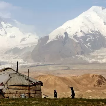 A felt yurt and two small figures on a grassy rise, with glaciers and snow-capped mountains behind.