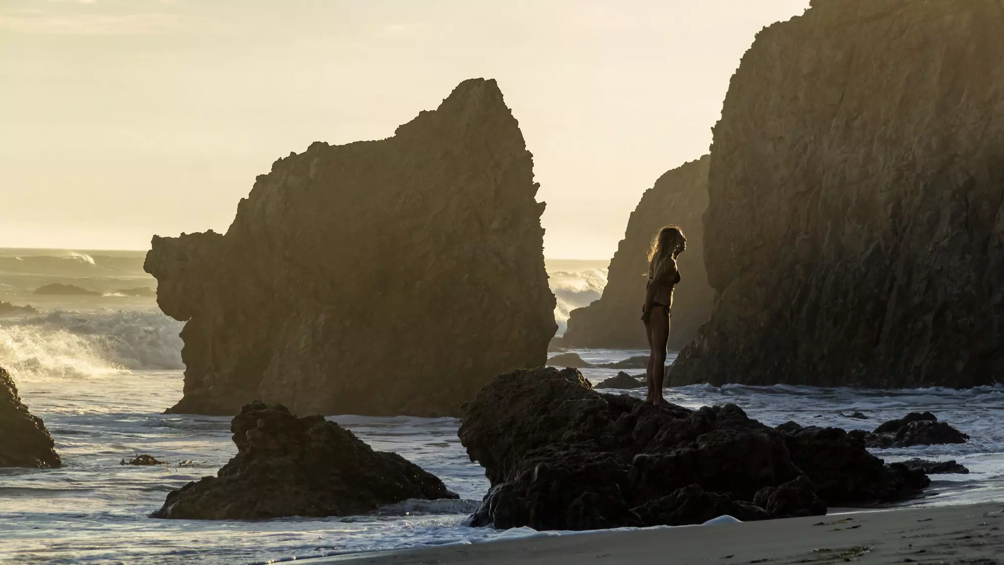 A girl stands on a stone cliff on El Matador Beach at sunset on a clear sunny day. Concept, leisure, tourism, travel.