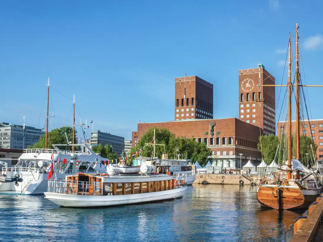 Red-brick city hall with two towers, one bearing a large clock, behind moored excursion boats and a wooden two-masted ship