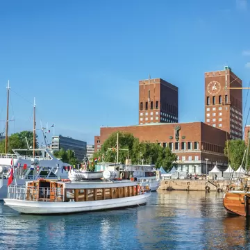 Red-brick city hall with two towers, one bearing a large clock, behind moored excursion boats and a wooden two-masted ship
