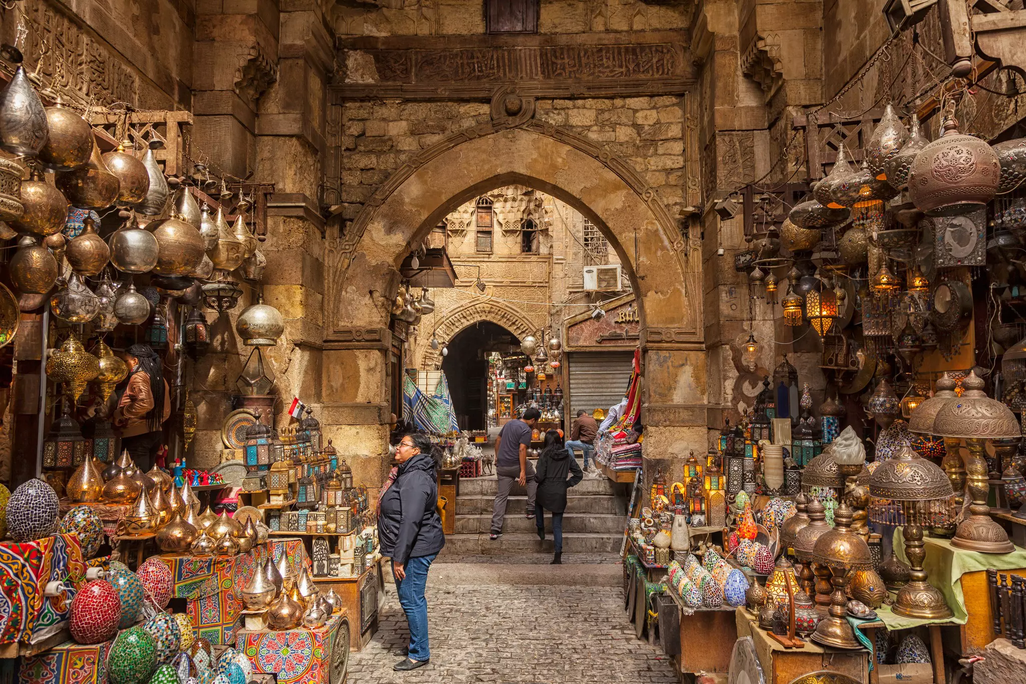 A shopper looks at lanterns hanging from the wall and stacked on shelves at an outdoor stand by an archway on a street in Cairo, Egypt.