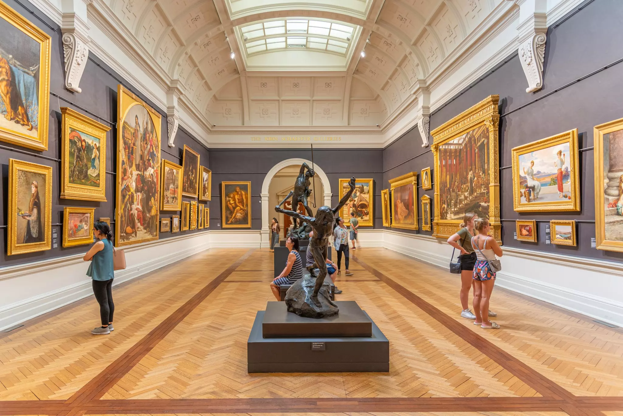 An art gallery hall with walls lined with works of various sizes. Sculptures are in the middle of the hall.