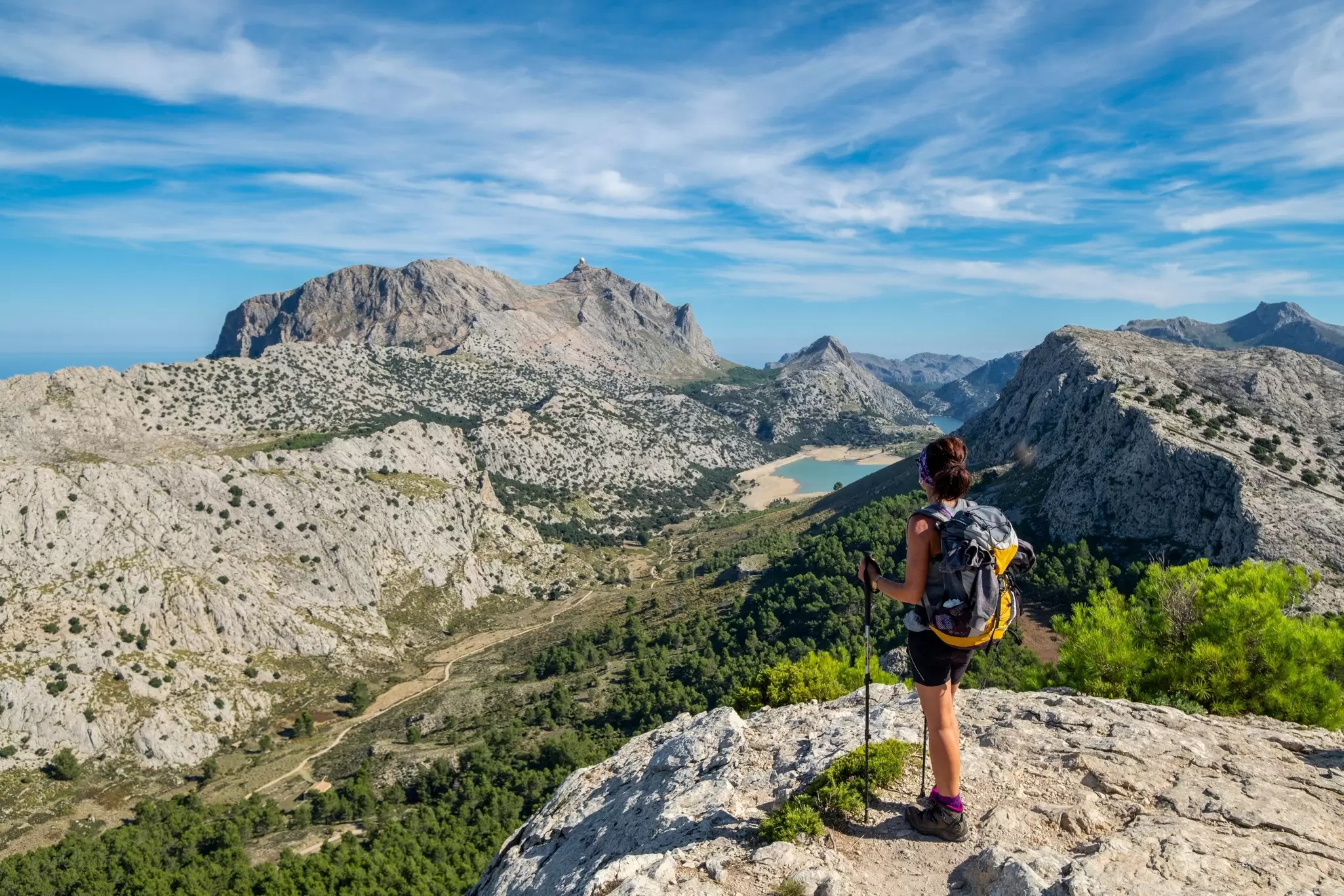 Hiker with a backpack and trekking poles stands on a rocky ridge overlooking rugged peaks and a turquoise reservoir