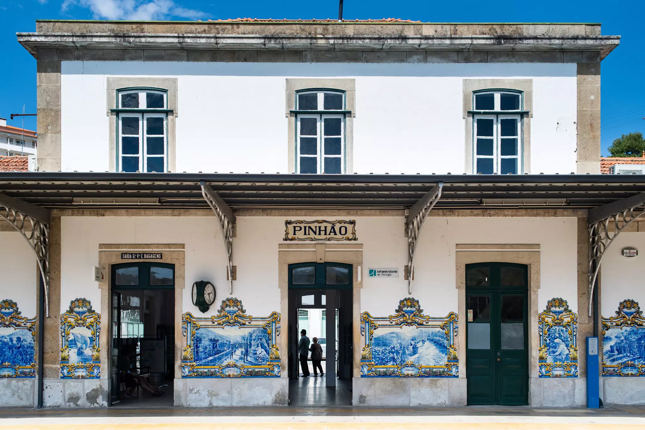 A two-story white train station with blue and white painted tiles and a sign that says Pinhão.