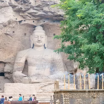 A giant stone Buddha carved into a sandstone cliff, with a leafy tree to one side and visitors at a railing below.