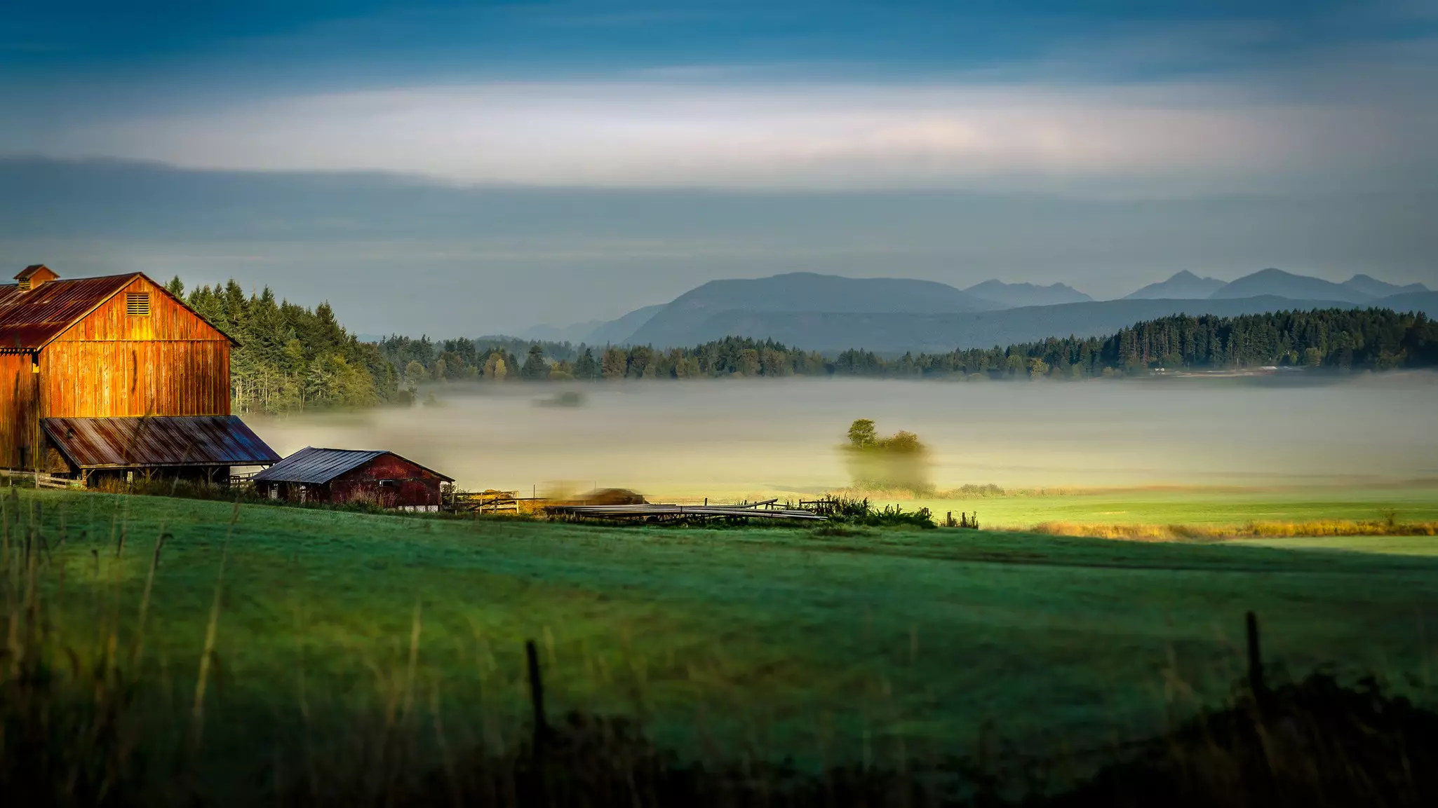 Mist hangs over a green valley; sunlight hits a wooden structure.