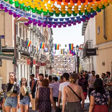 Shoppers on a sunny street in Madrid's Chueca under rainbow garlands of red, yellow, green and purple cups and Pride flags.