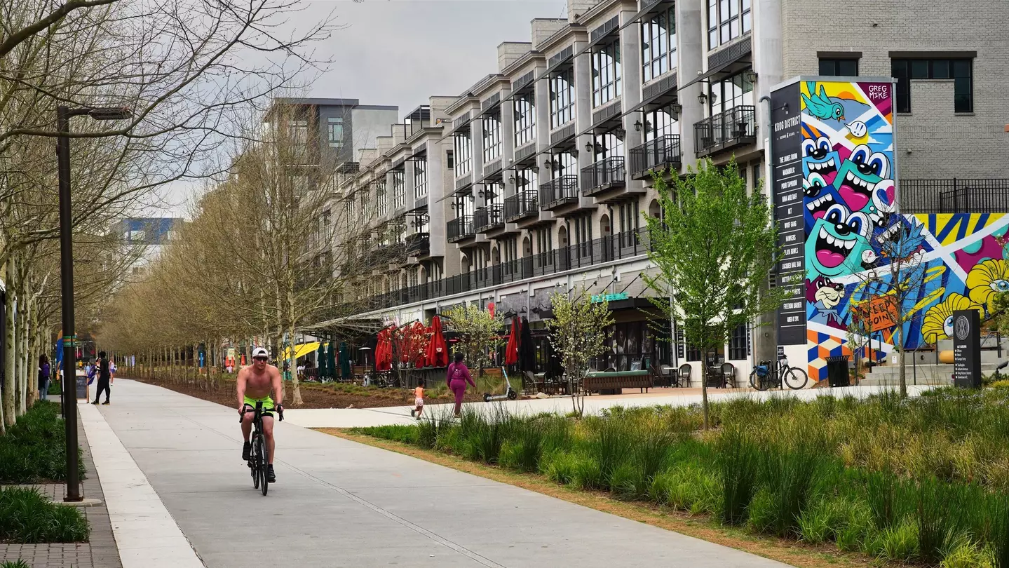 A cyclist pedals down a path next to trees and apartment complexes in a city.