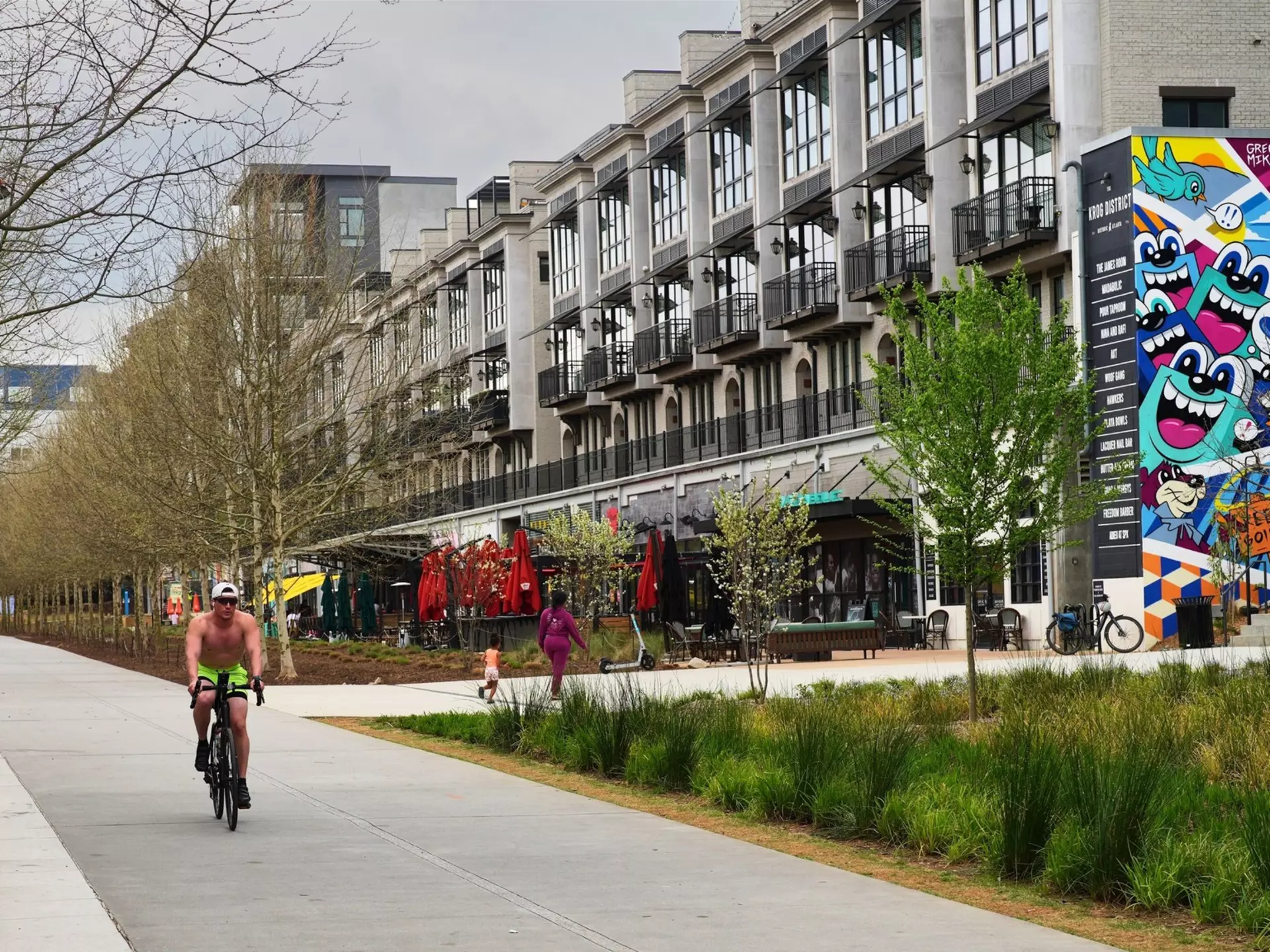 A cyclist pedals down a path next to trees and apartment complexes in a city.