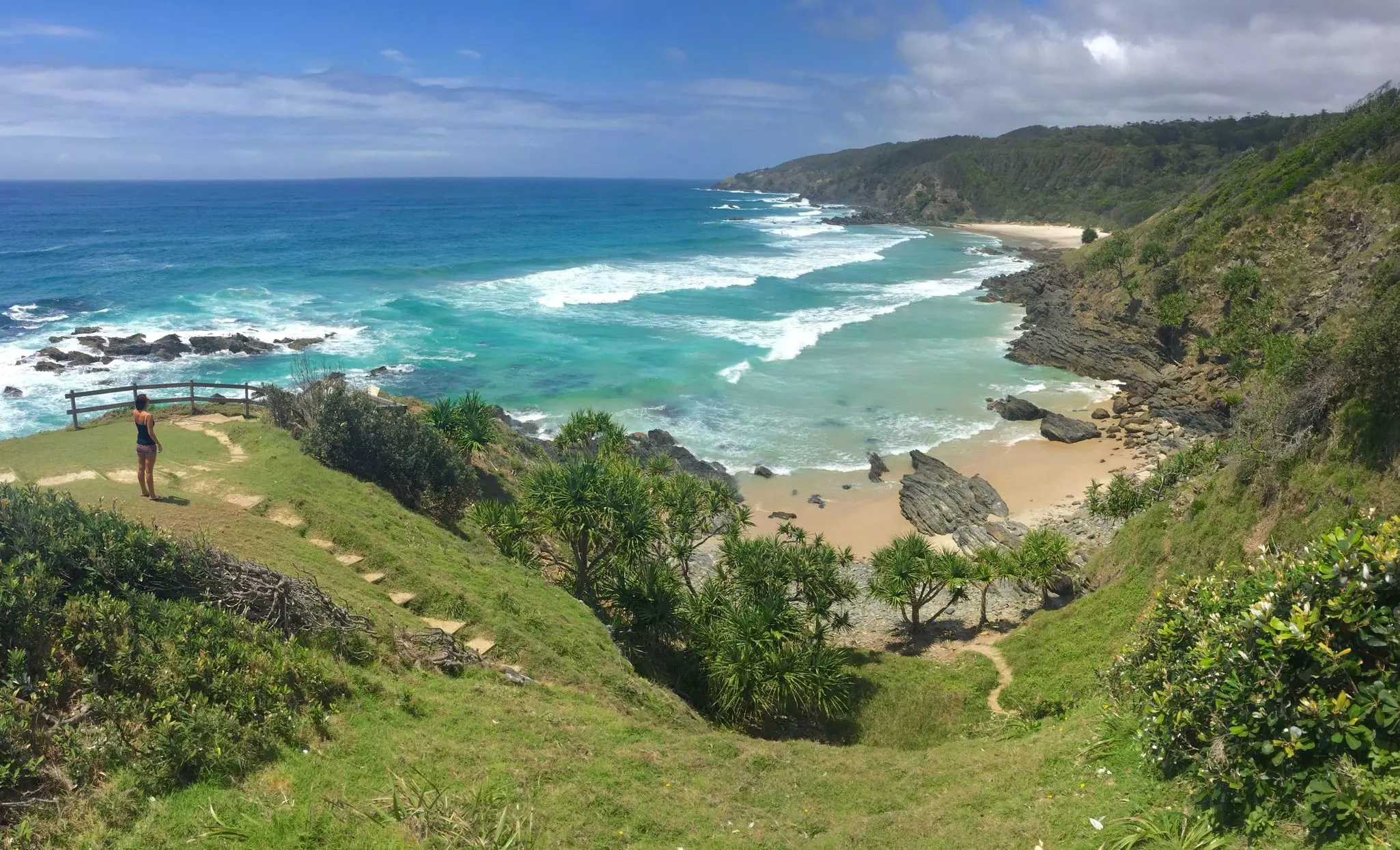 A person stands on a grassy hill overlooking a the ocean and a small beach