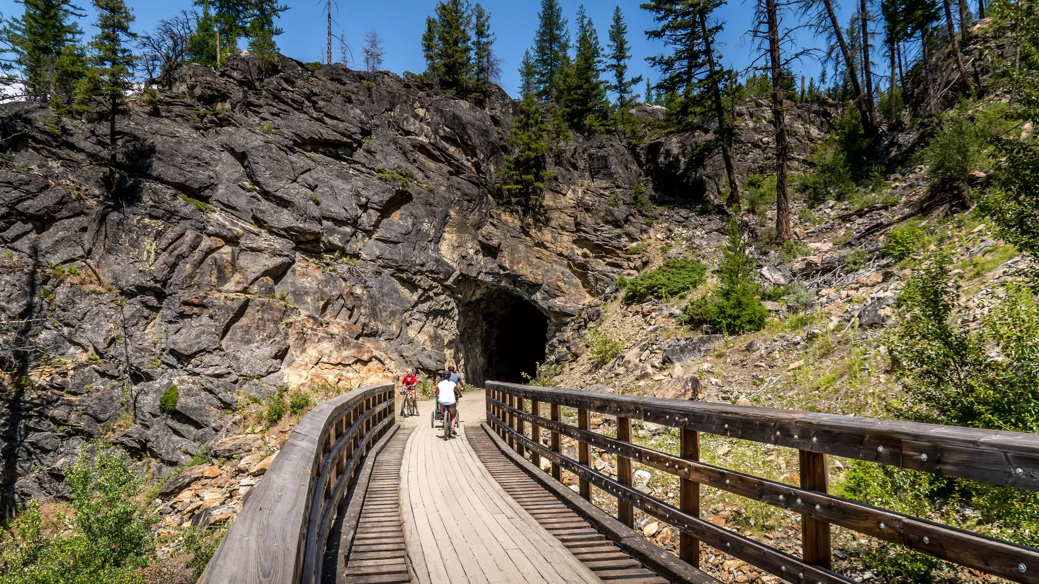 A cyclist pedals over  wooden trestle bridge heading towards a tunnel through rock on a former rail line.