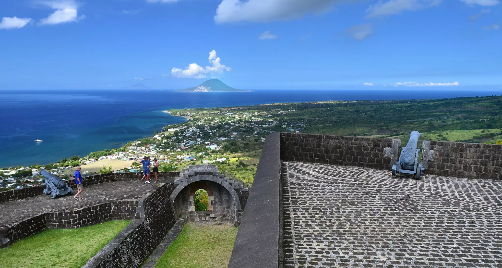 Tourists wander around a historic hilltop fortress with high walls and canons pointing out to sea.