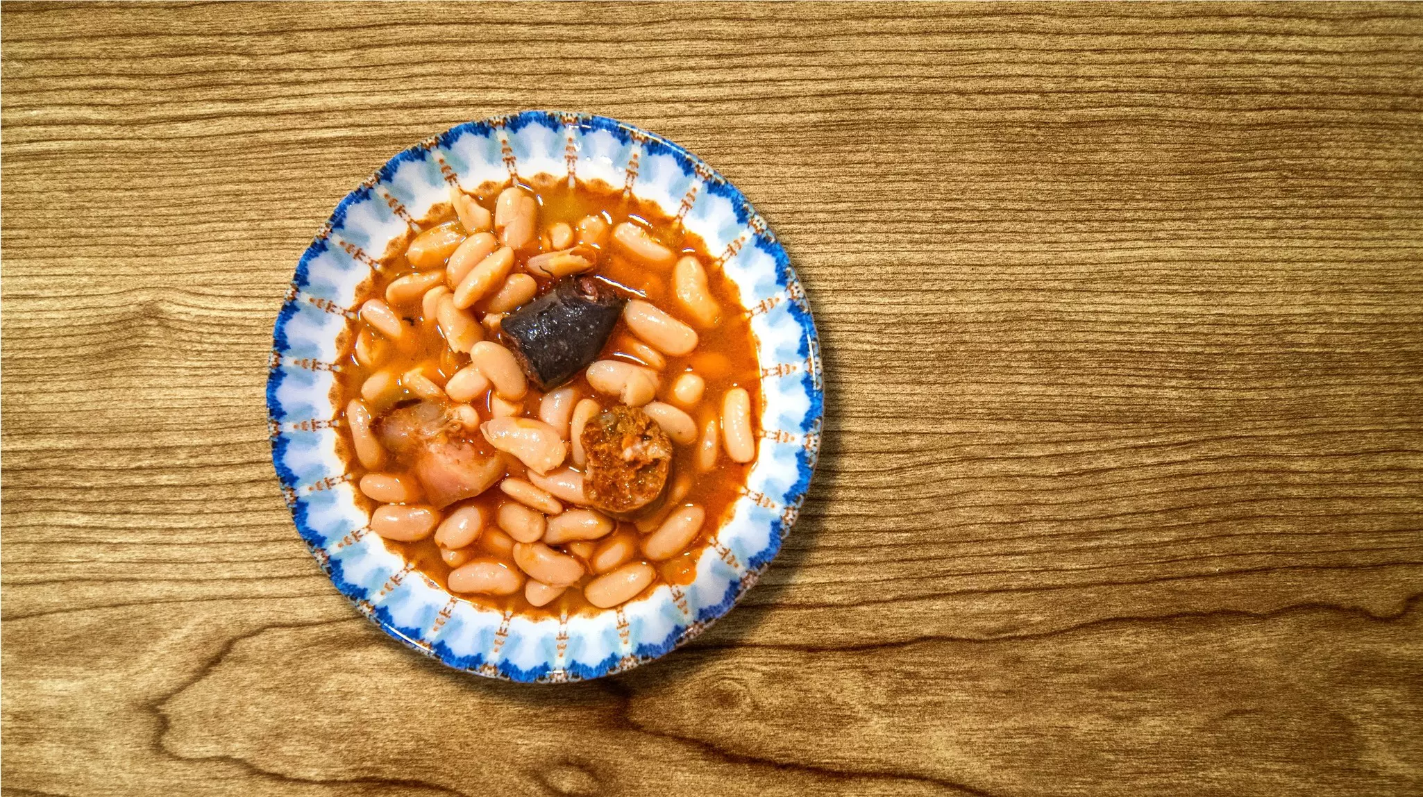 On a wooden table sits a bowl filled with a stew made from white beans, black pudding and chorizo.