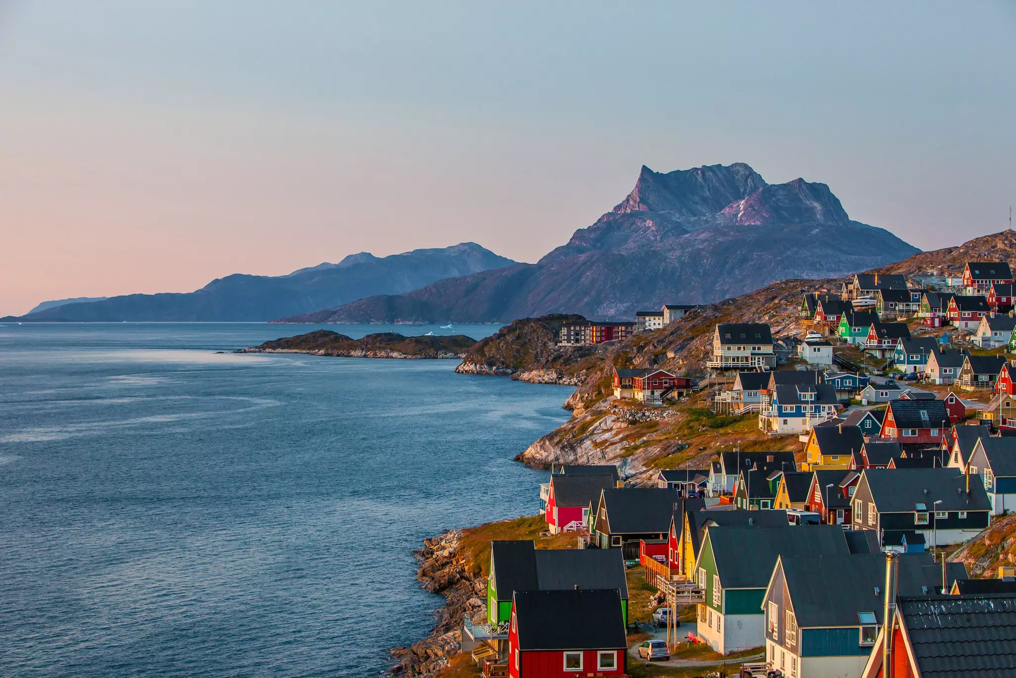 Evening light falls on buildings painted in red, blue and green, with steep peaked roofs, on a hillside by a body of water in Greenland; there are mountains in the background.