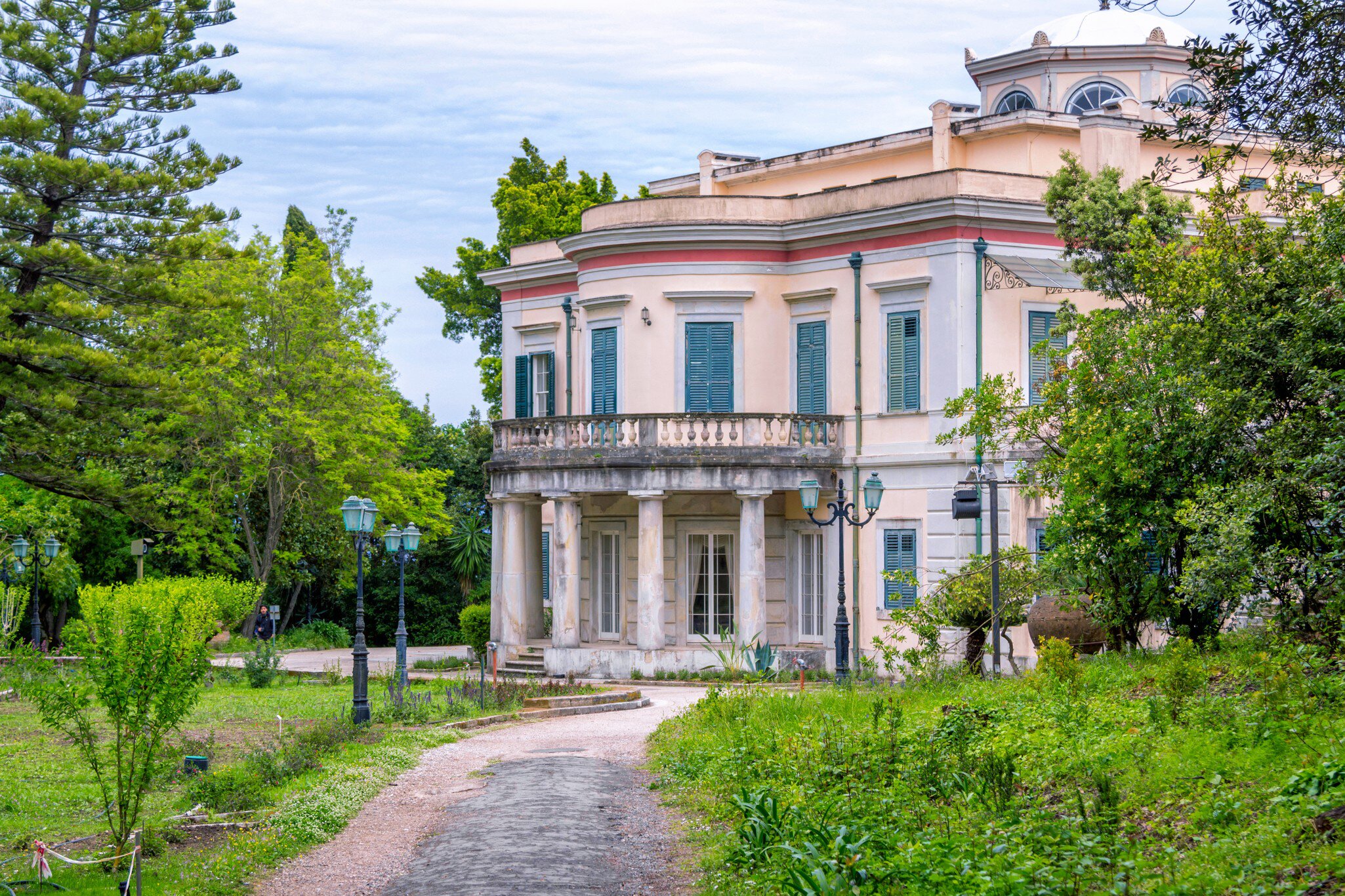 A pale neoclassical villa with green shutters and a balustraded balcony on marble columns, at the end of a gravel path through overgrown gardens.