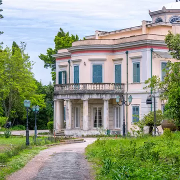 A pale neoclassical villa with green shutters and a balustraded balcony on marble columns, at the end of a gravel path through overgrown gardens.