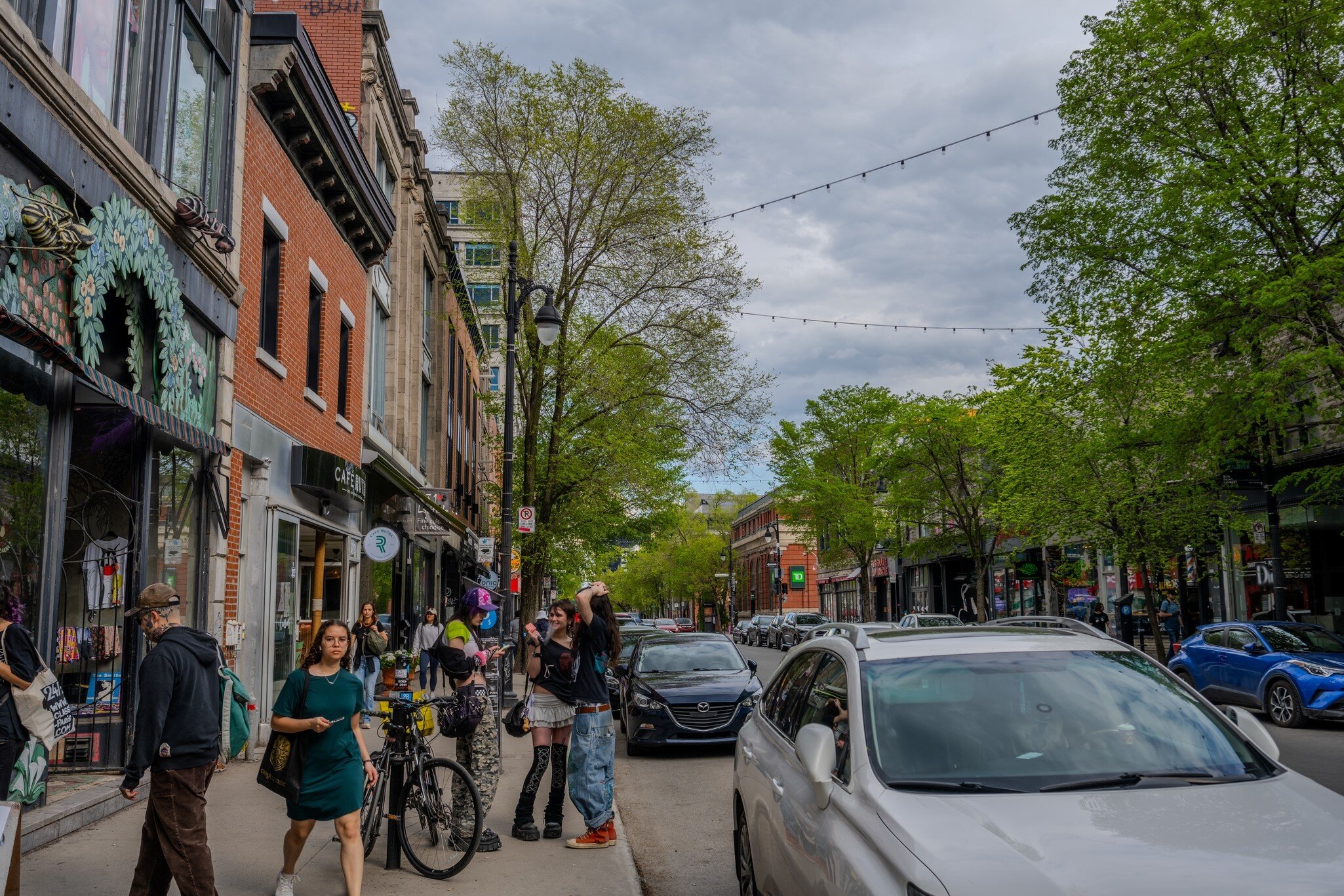 Pedestrians walk past shops and parked cars on Saint Laurent Boulevard in Montreal under overcast skies and spring foliage. 