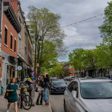 Pedestrians walk past shops and parked cars on Saint Laurent Boulevard in Montreal under overcast skies and spring foliage. 