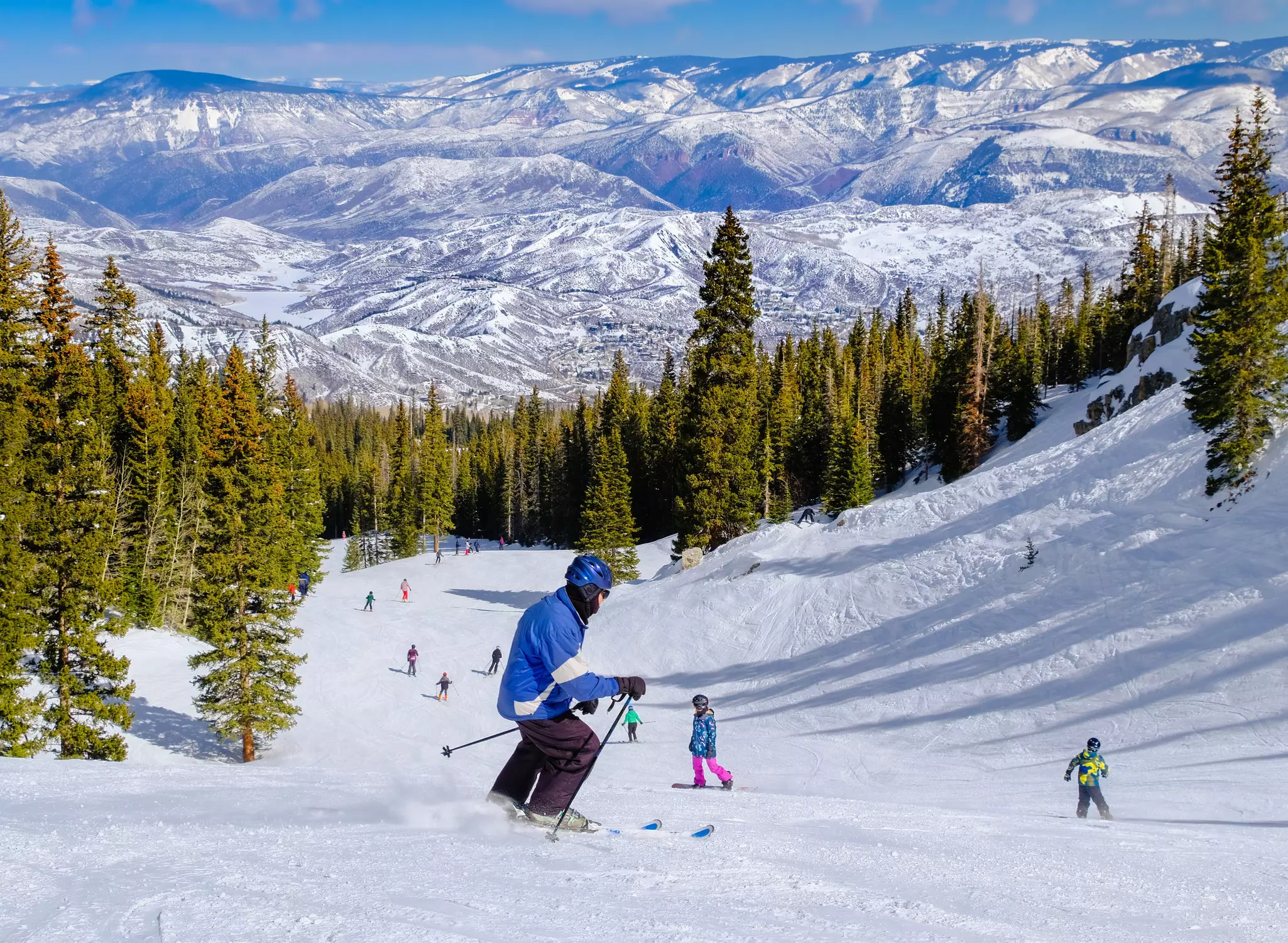 A skier going downhill toward other skiers, with views of the mountains and alpine trees.