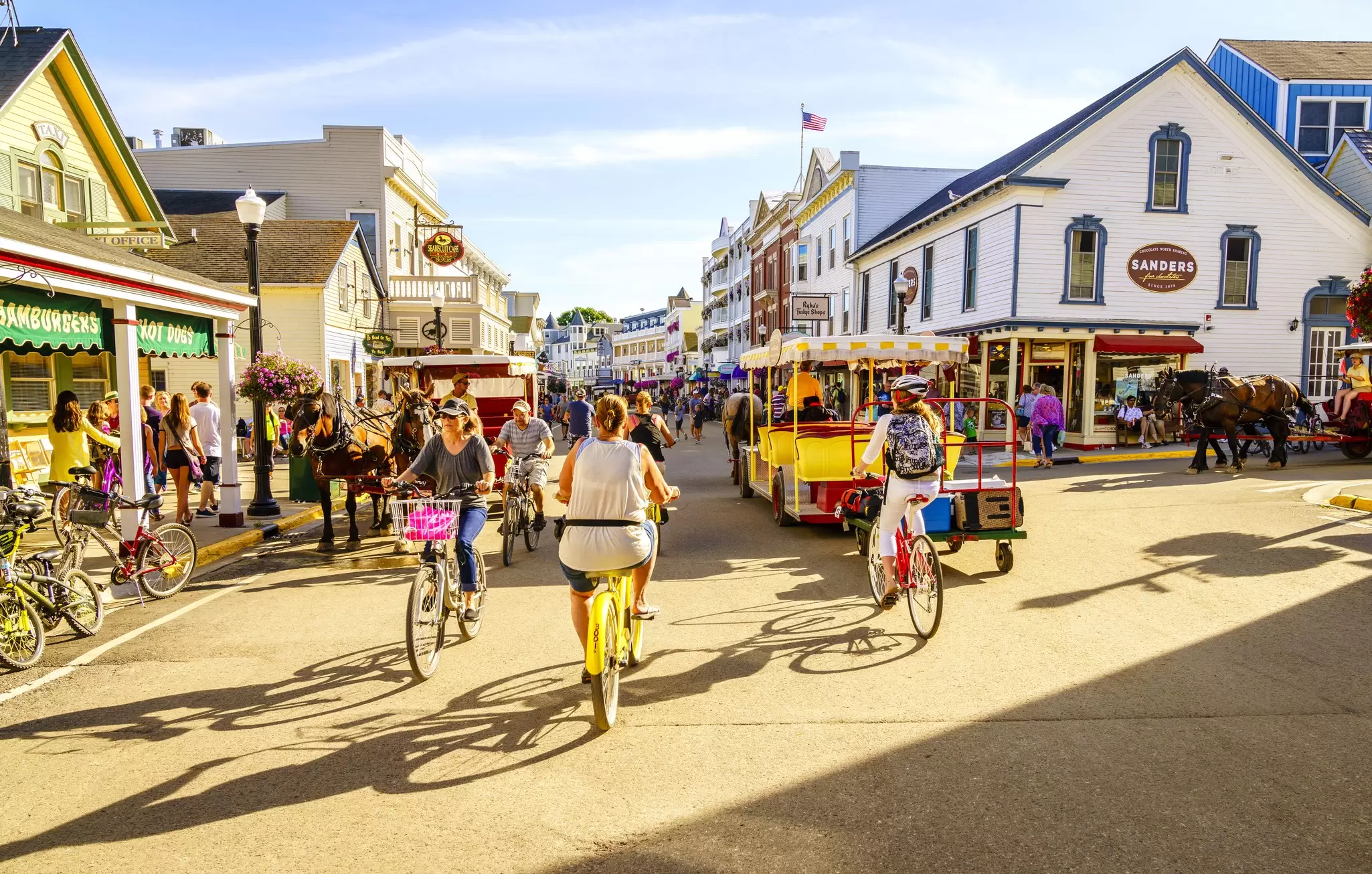 People walk and bicycle on a crowded street lined with flowerpots and storefronts.