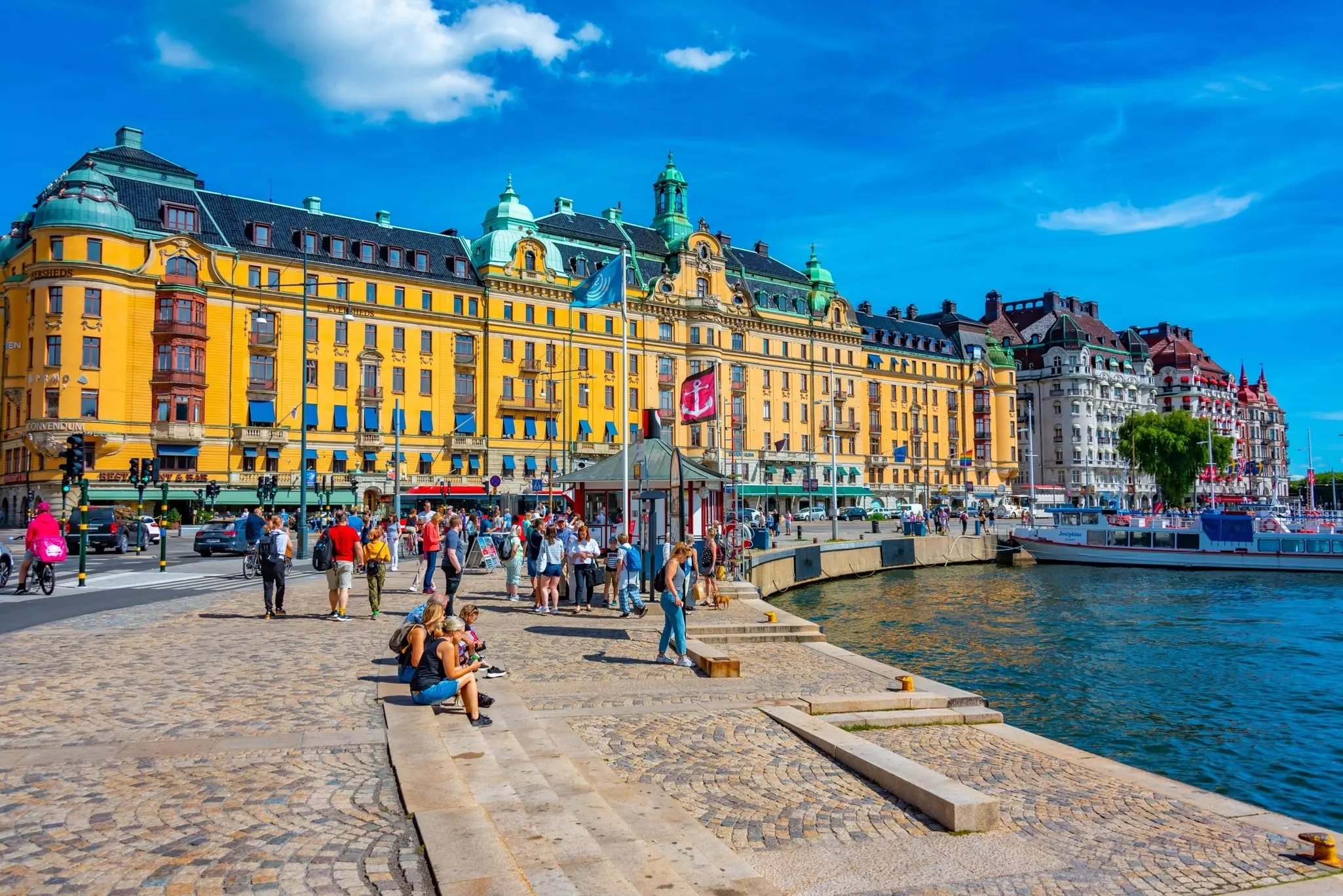 People walk along a waterfront promenade in a city, in front of a grand building painted yellow.
