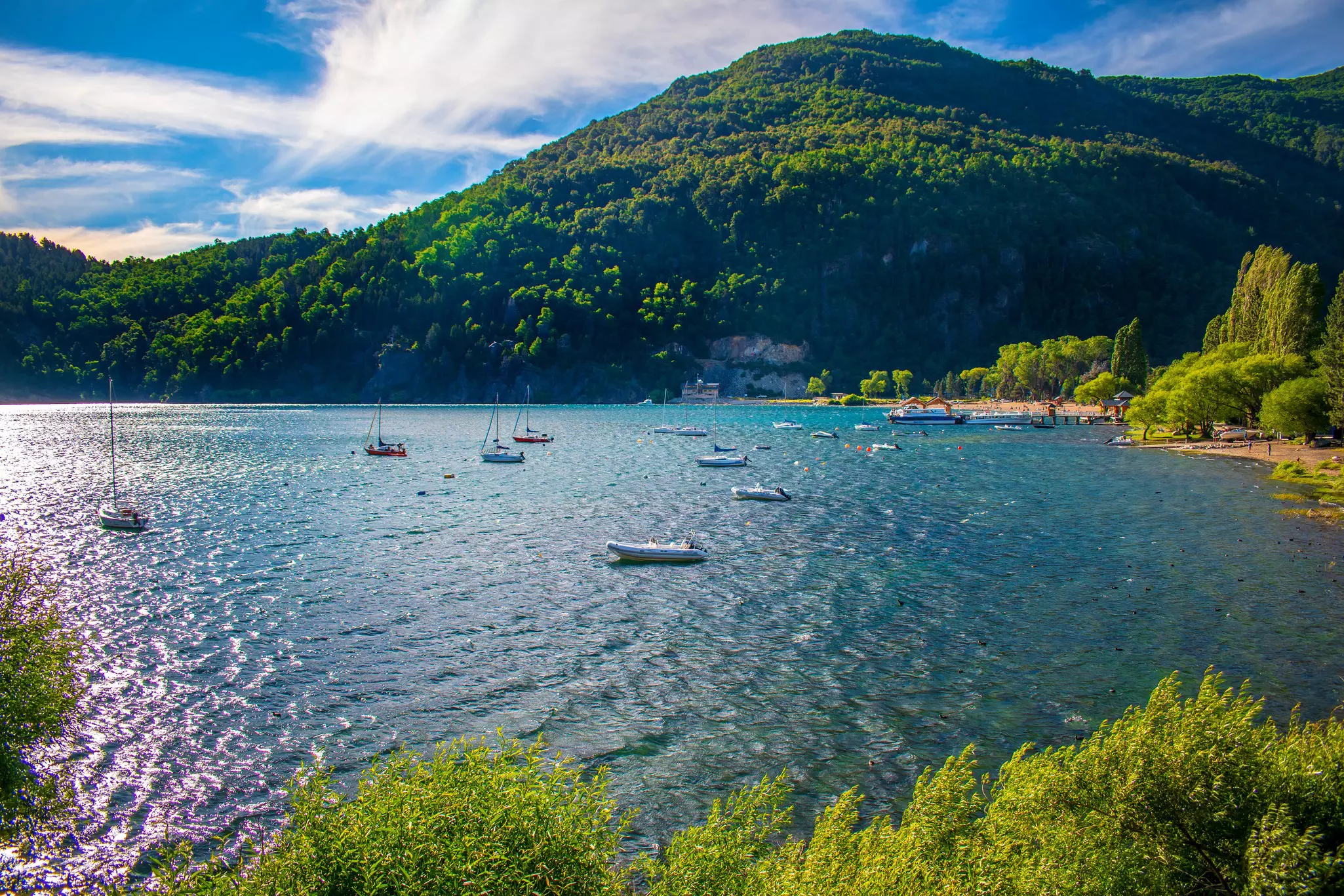 A lake with small boats on a sunny day. People relax on the sandy beach.