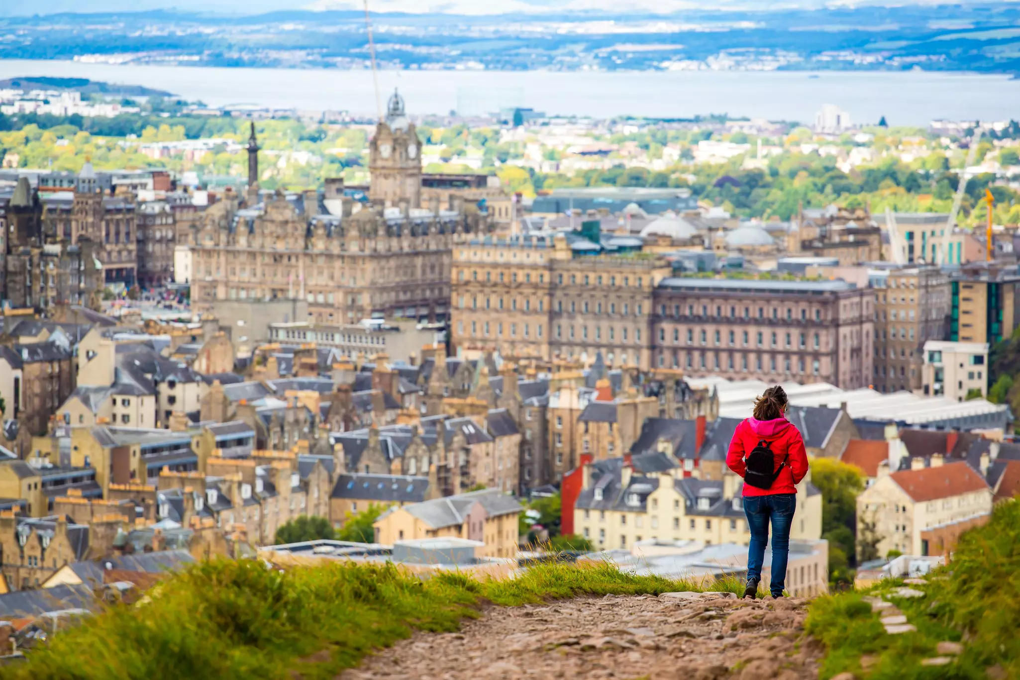 Edinburgh seen from the trail to the top of Arthur's Seat, with a hiker in a red coat, Edinburgh, Scotland.