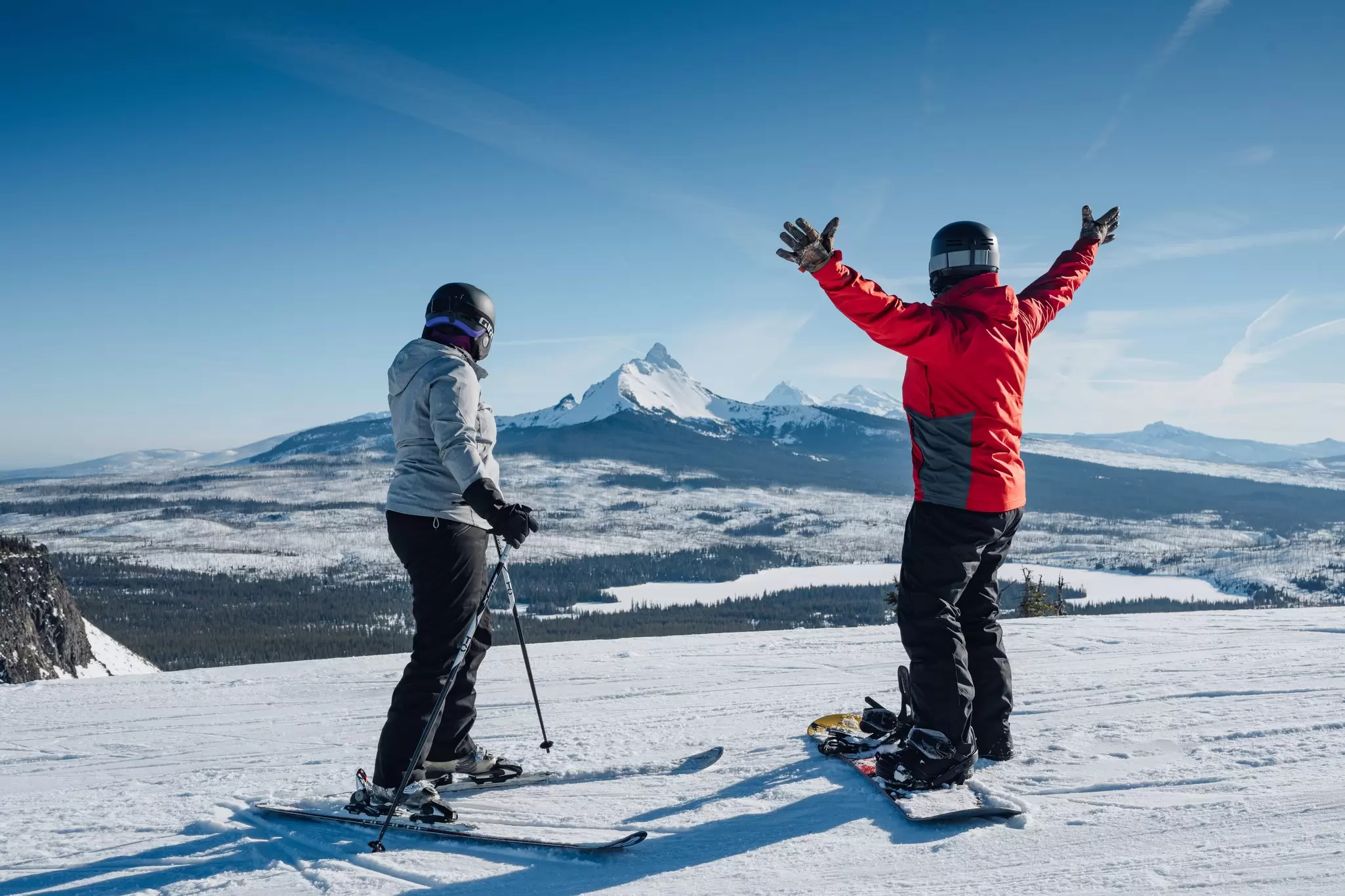 One woman on skis another on a snowboard with arms up looking at the snowy mountain landscape in Hoodoo Ski Area in Sisters, OR
