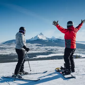 One woman on skis another on a snowboard with arms up looking at the snowy mountain landscape in Hoodoo Ski Area in Sisters, OR

