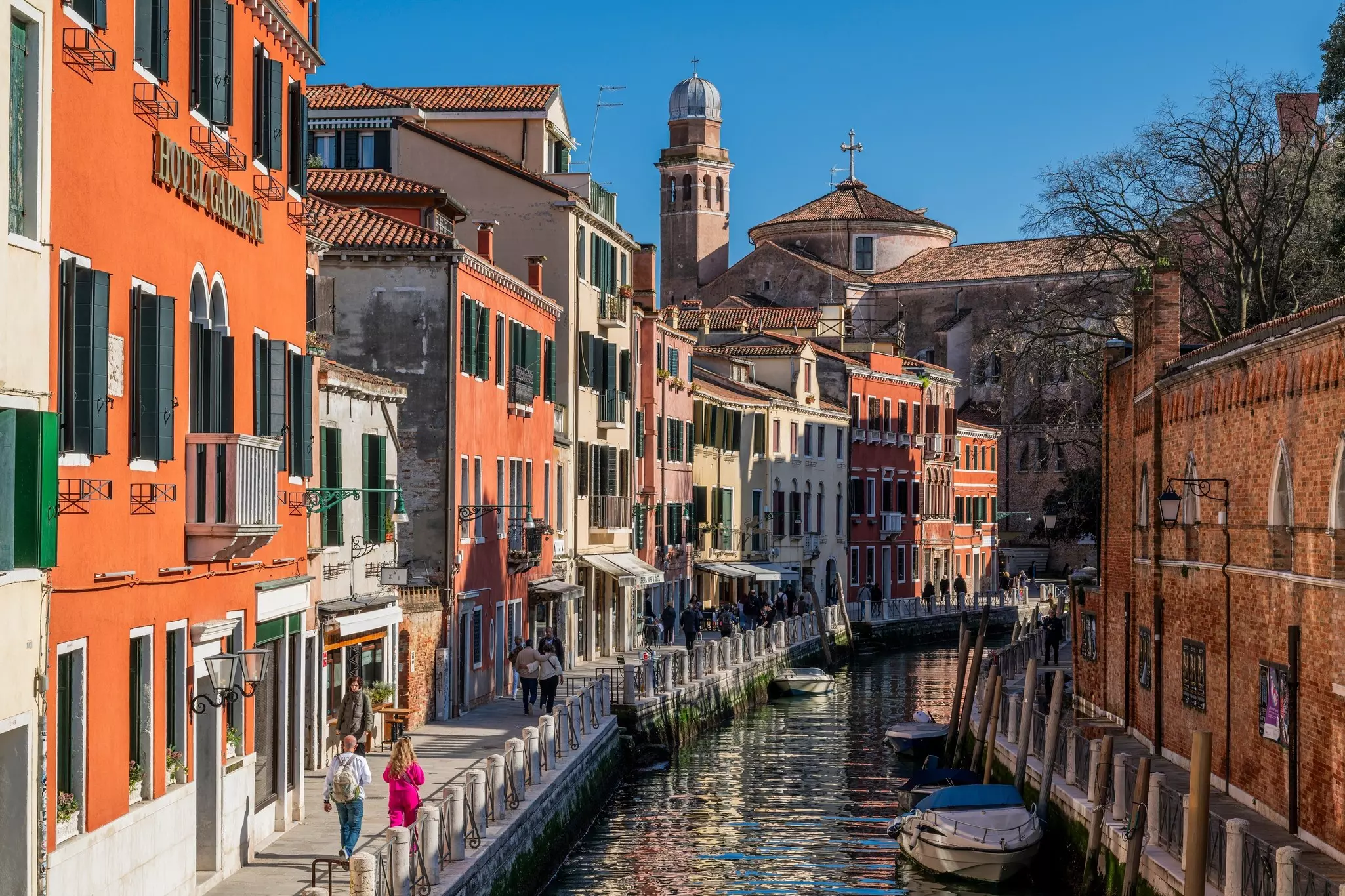 People walk along the waterfront in Venice on a street with terracotta-colored buildings