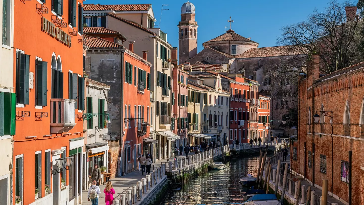 People walk along the waterfront in Venice on a street with terracotta-colored buildings