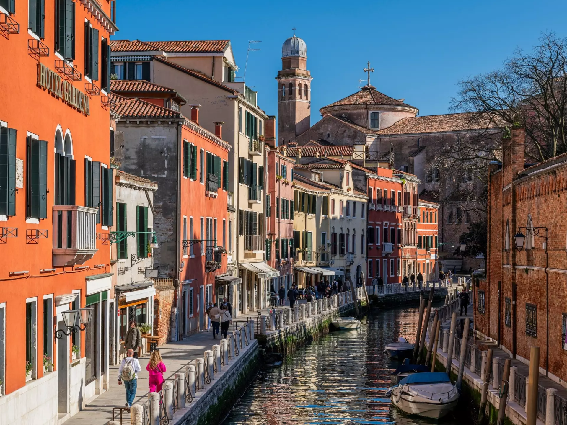 People walk along the waterfront in Venice on a street with terracotta-colored buildings