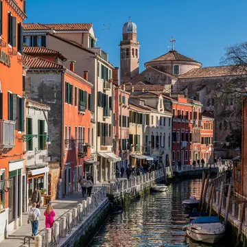 People walk along the waterfront in Venice on a street with terracotta-colored buildings