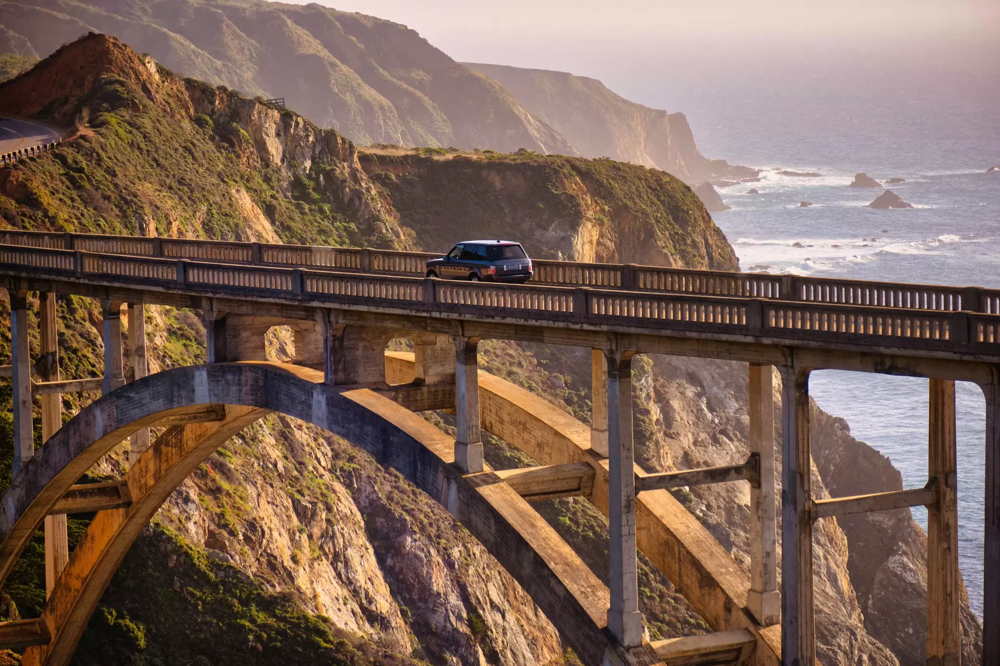A car drives on a bridge over a ravine by the coast at sunset.