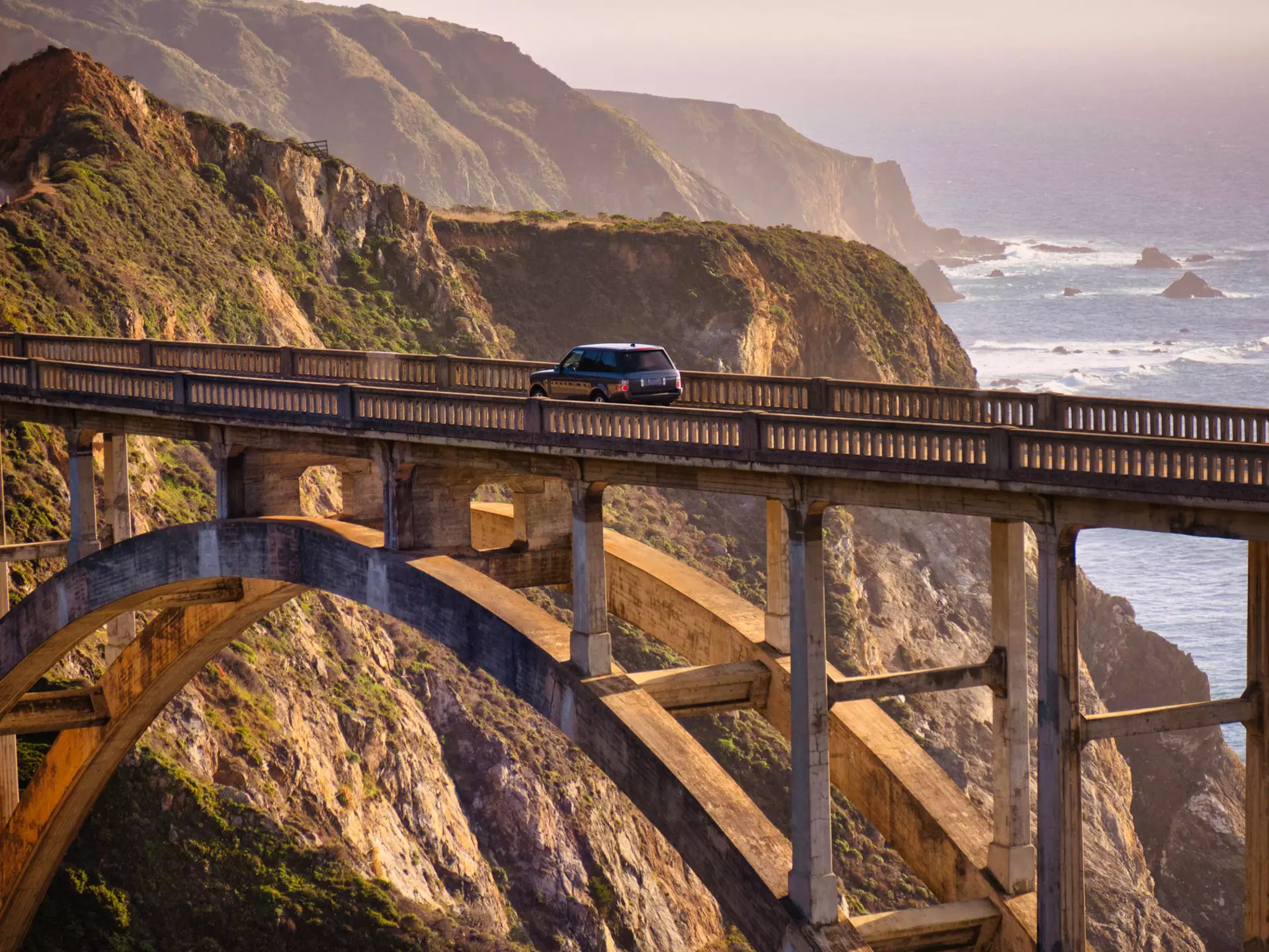 A car drives on a bridge over a ravine by the coast at sunset.