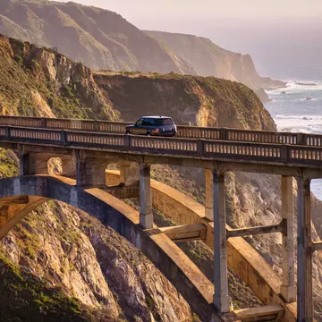 A car drives on a bridge over a ravine by the coast at sunset.