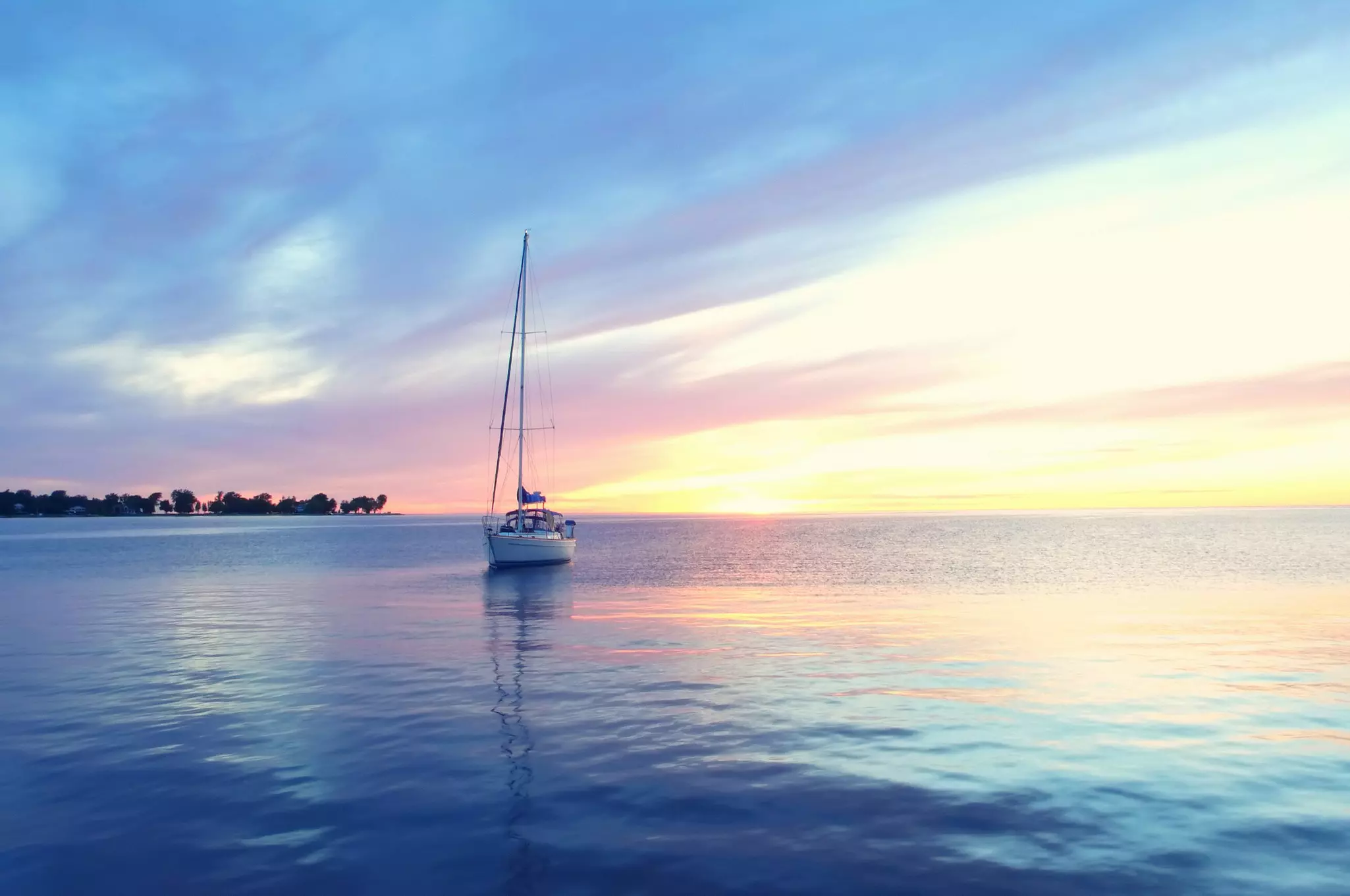 A single-sail boat anchored in calm waters as the sunset casts pastel colors across the sky