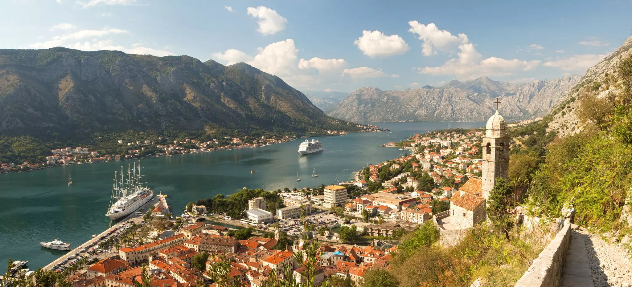 An aerial view of a town in Montenegro on a hillside by a wide body of water; there are large boats in the water.