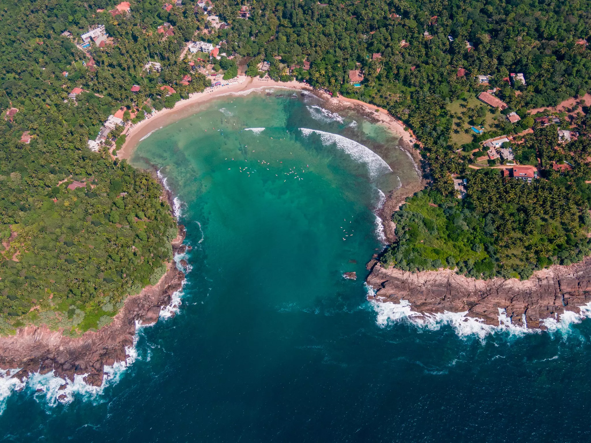 An aerial view of a horseshoe-shaped cove with a beach and houses, framed by two rocky, tree-covered headlands.