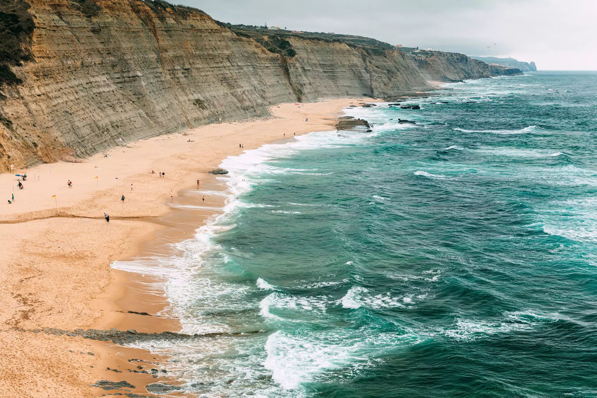 A wide view of white-capped waves washing ashore at a beach backed by tall cliffs, with about a dozen bathers visible
