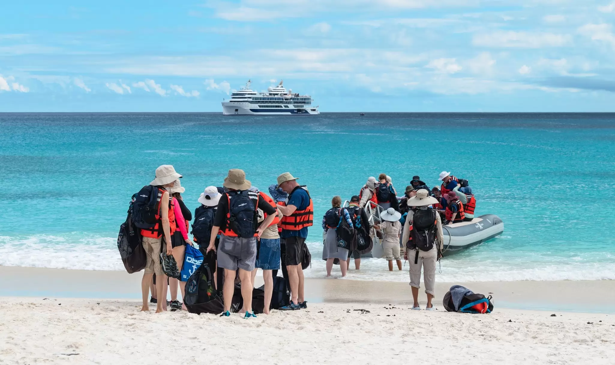 Visitors disembark from Zodiac tenders on a beach, their cruise ship visible in the waters offshore, Española Island, Galápagos Islands